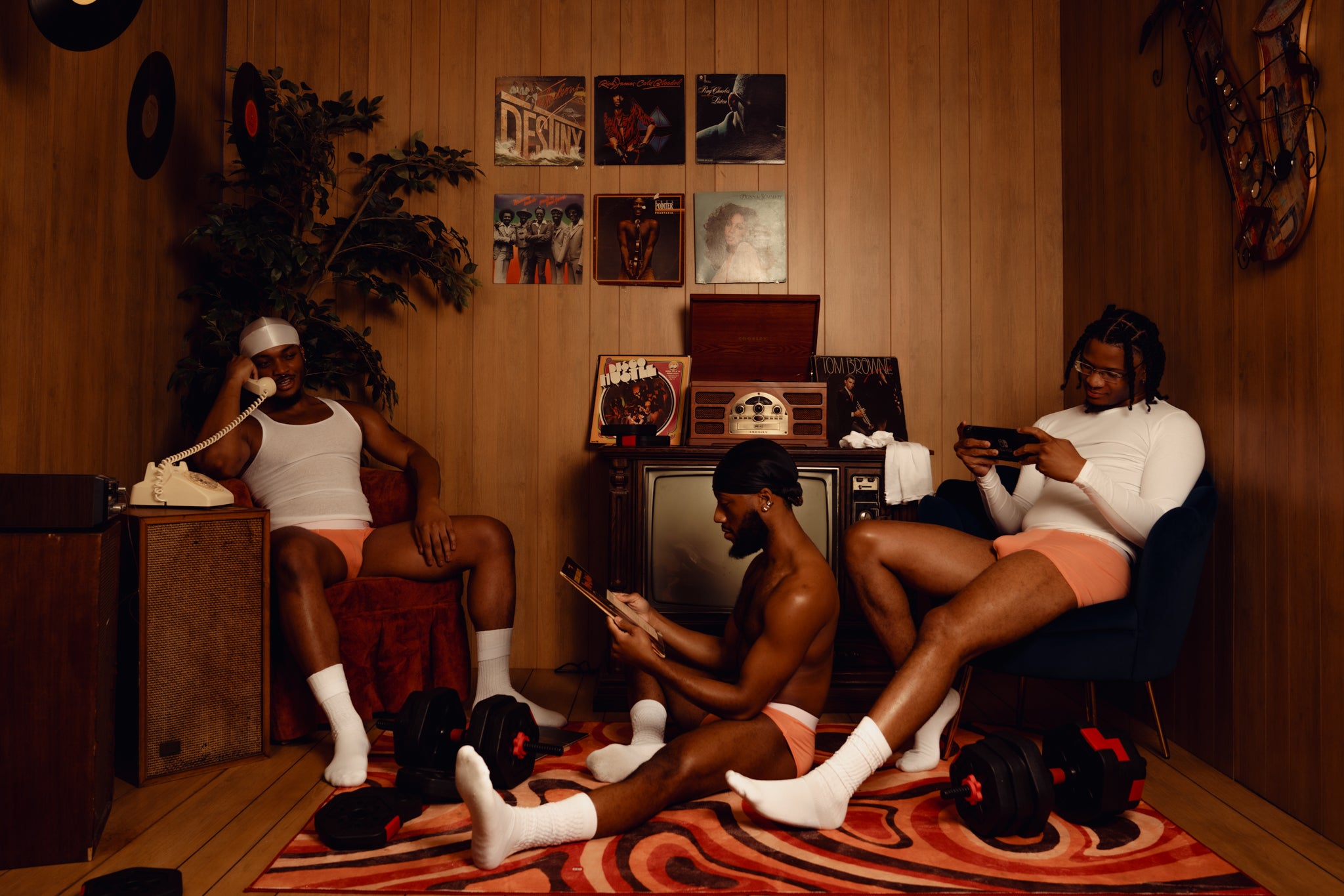 Three people sitting in a room with vinyl records, and a television modeling "AKATE Classic Peach Briefs & Boxers."