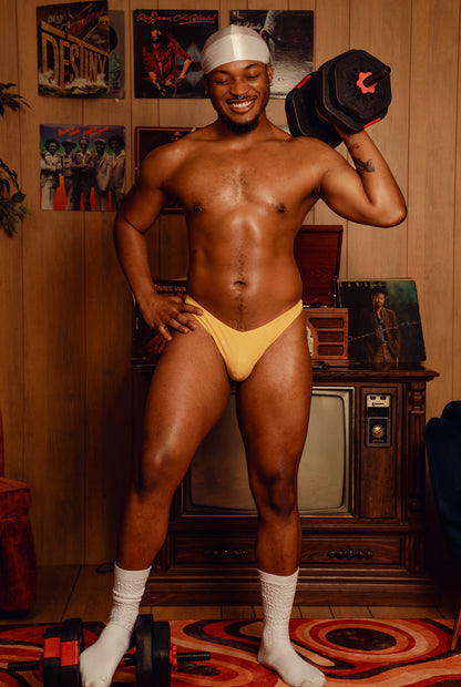 Man posing with weights in a room with vinyl records and a television sporting an Akate Classic Thong.