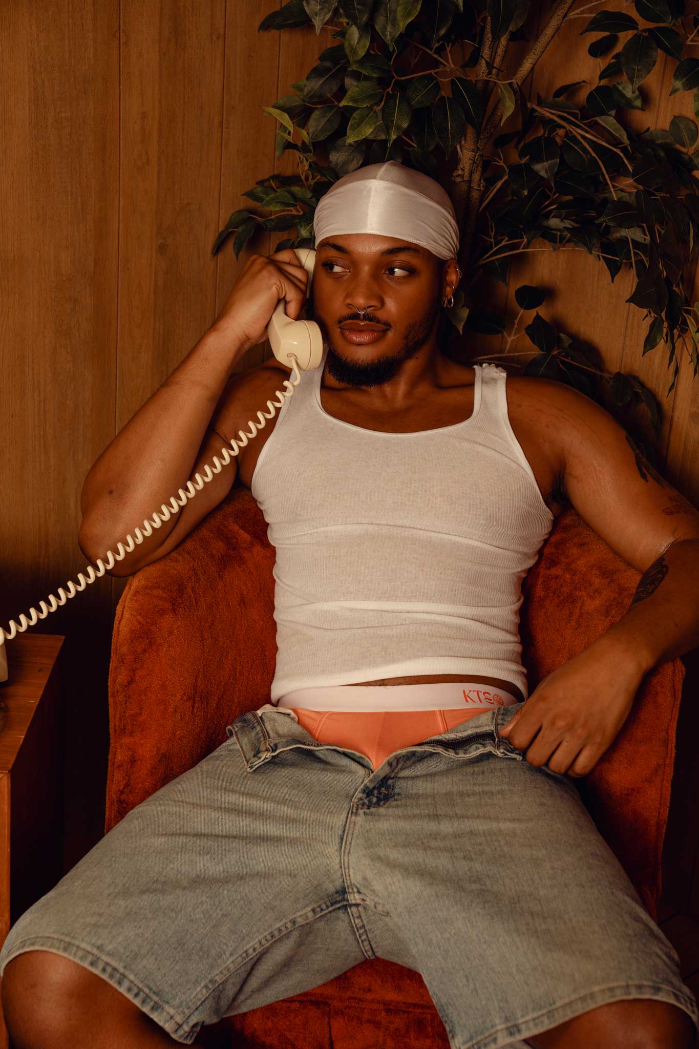 Man sitting on a couch wearing a white tank top, peach AKATE Classic Boxer Briefs (also coming in midnight black), & denim shorts, holding a phone to his ear.